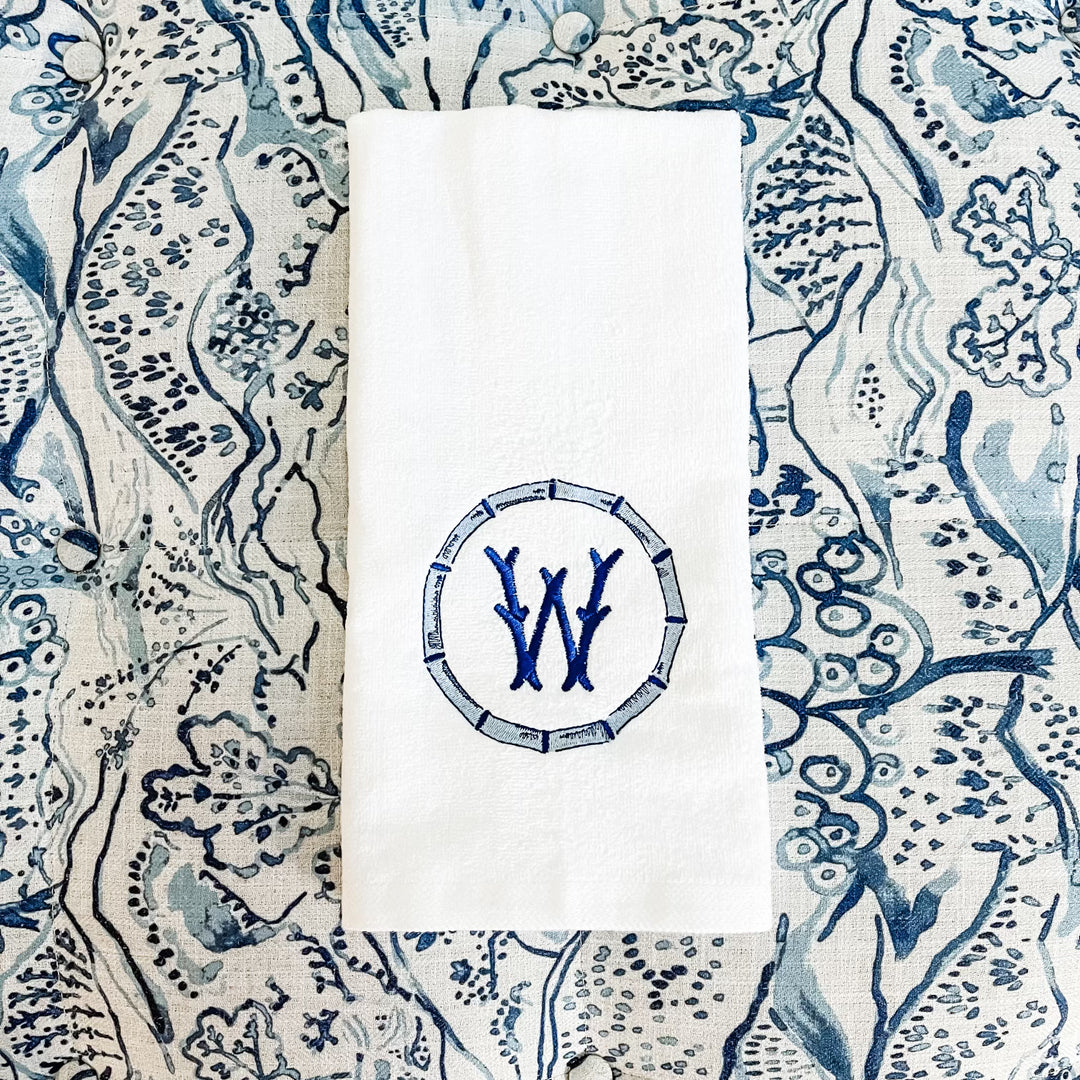 A white hand towel with a blue bamboo circle and a single initial monogram, placed on a patterned blue and white textile surface.
