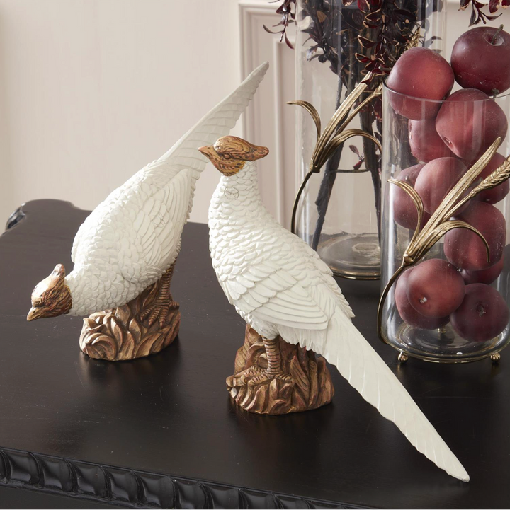 Decorative pheasant statues on a dark surface with a glass container of fruits in the background.