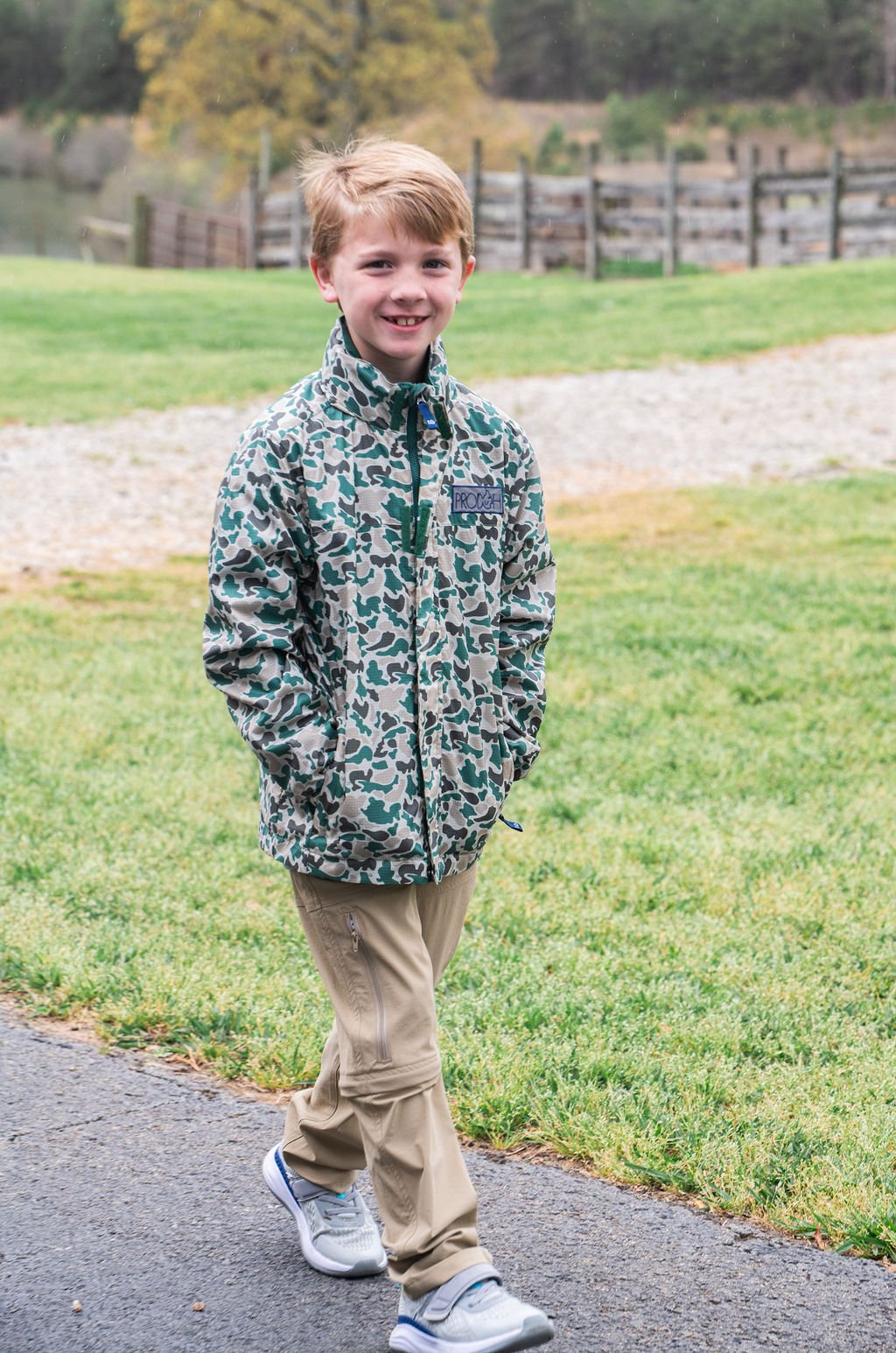 Young boy wearing a green camouflage jacket standing on a path with grass and trees in the background