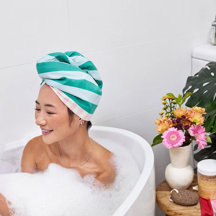 Woman in a bubble bath with a striped towel on her head, surrounded by flowers and a plant.