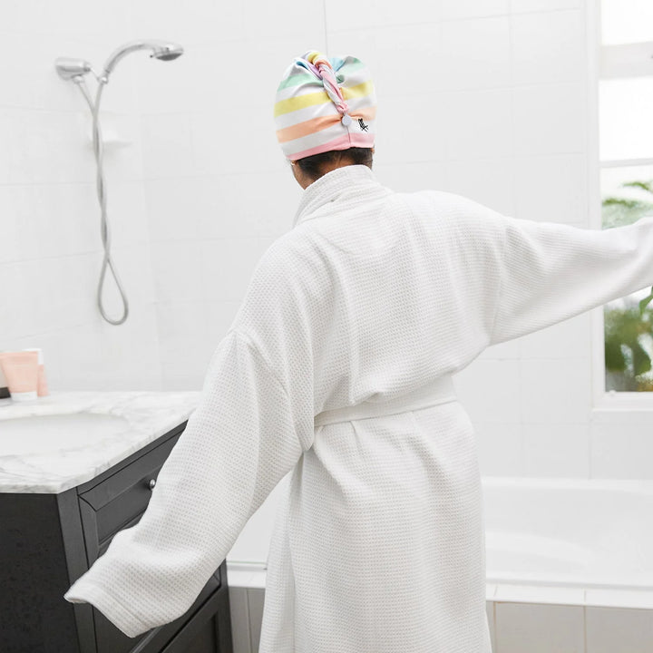 Person wearing a colorful striped towel on their head and a white robe in a bathroom.