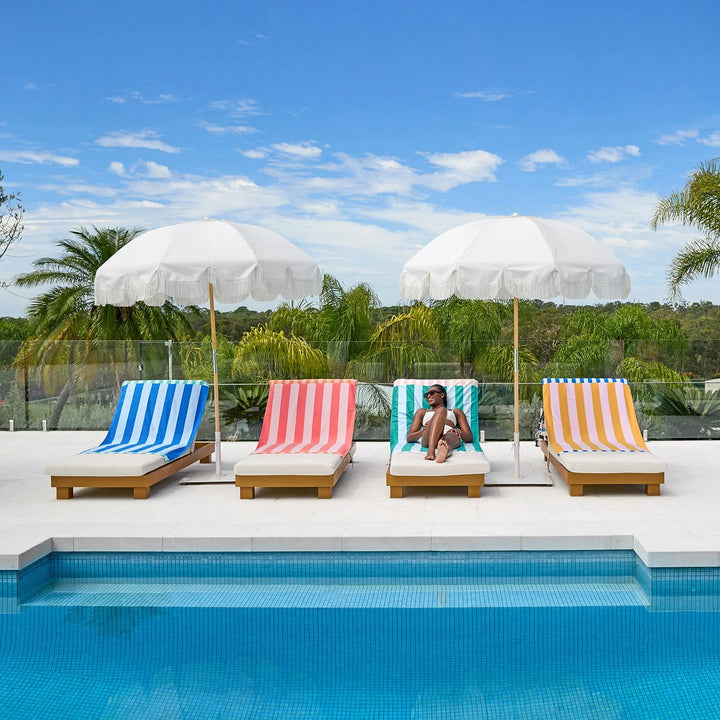 Four colorful lounge chairs with striped covers by a pool, each under a white umbrella.