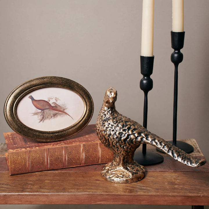 Decorative setup with a bronze pheasant figurine, framed picture, and candlesticks on a wooden surface.