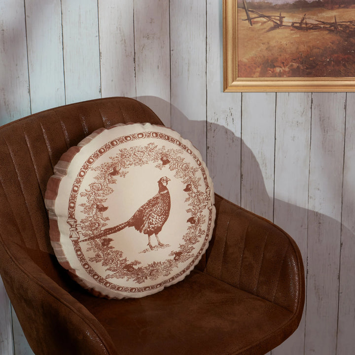 Round decorative pillow with a pheasant design on a brown chair against a wooden wall.