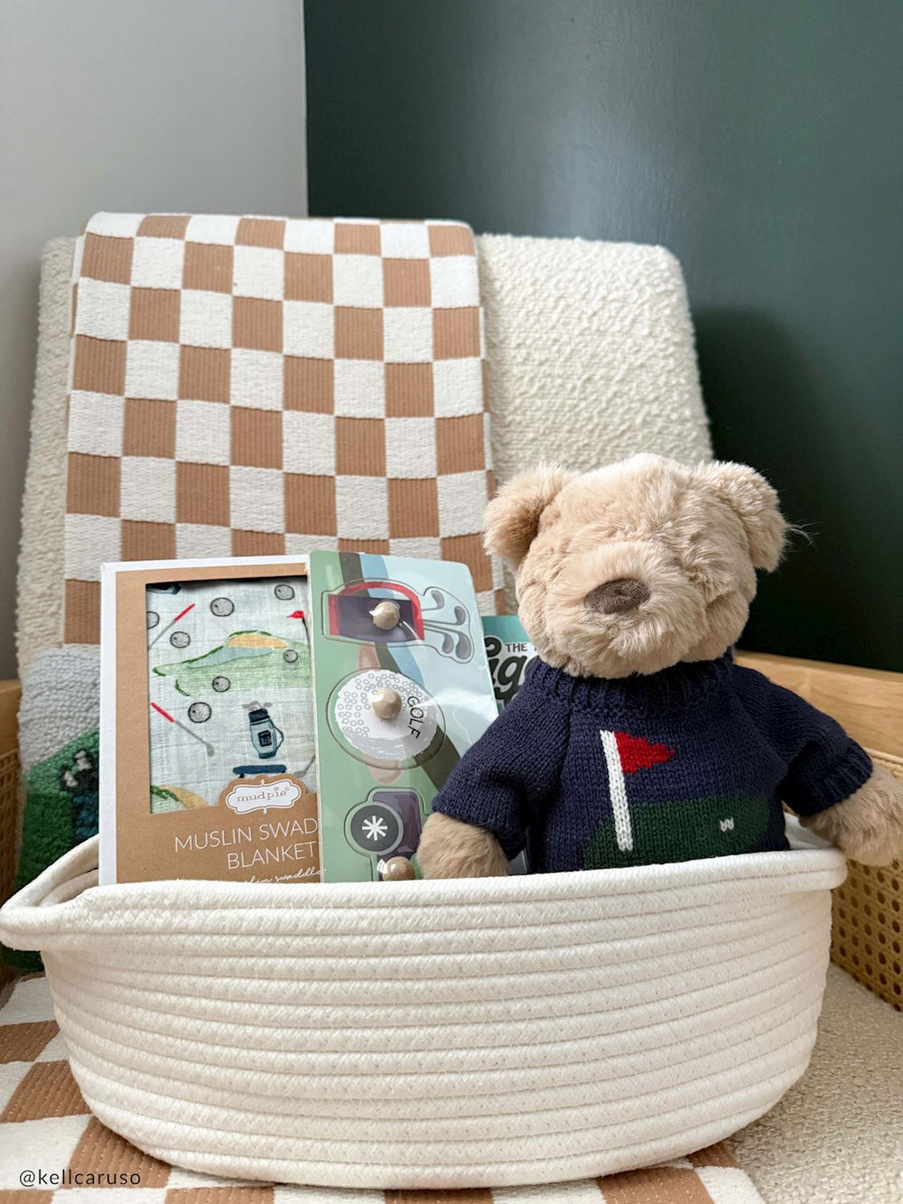 A plush teddy bear wearing a navy blue sweater sits in a cream rope basket next to a framed 'Muslin Swade Blanket' print, with a checkered blanket draped in the background.