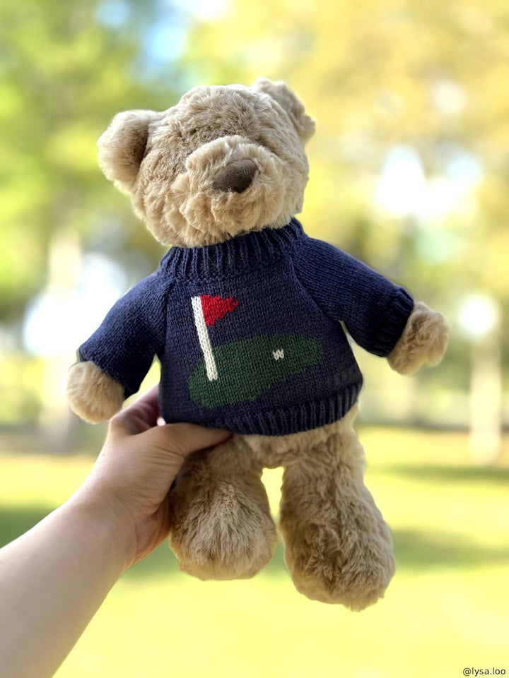 A plush teddy bear with light tan fur wearing a navy blue knit sweater with a green and white golf ball design on the chest, set against an outdoor background
