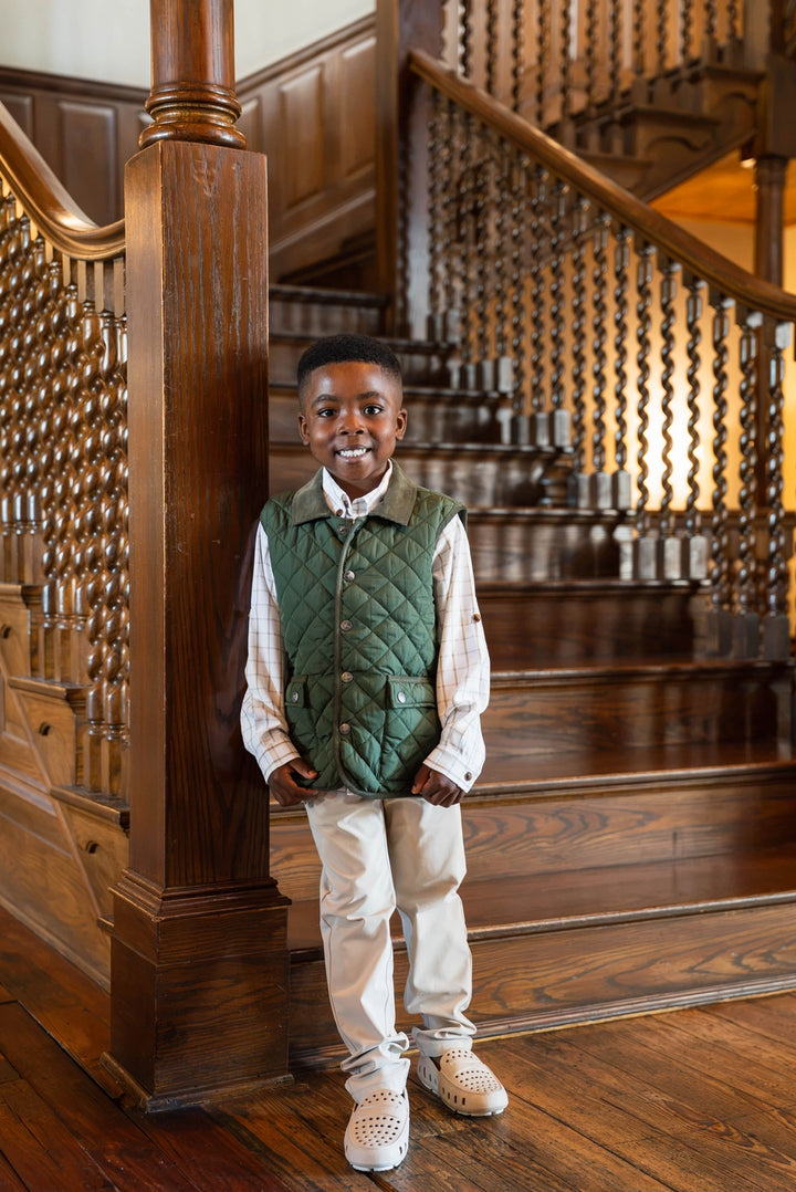 little boy wearing green quilted vest in front of wooden staircase