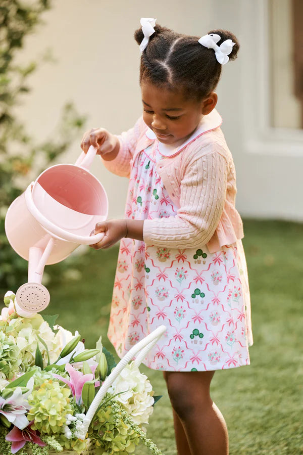Young girl in a floral dress holding a pink watering can and basket of flowers outdoors.