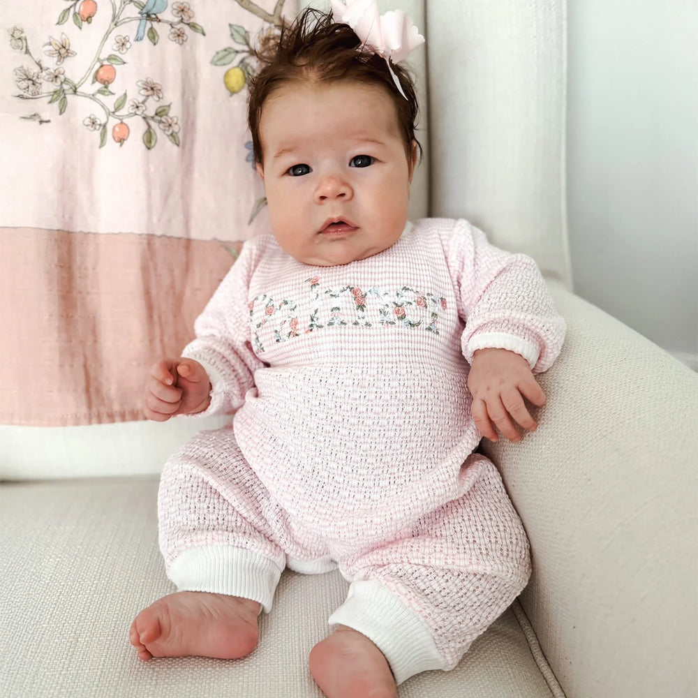 Baby in a pink knitted outfit sitting on a light-colored surface with a floral blanket in the background.