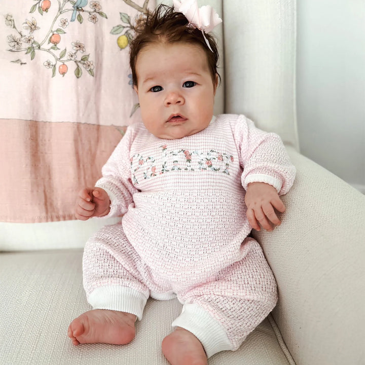 Baby in a pink knitted outfit sitting on a light-colored surface with a floral blanket in the background.