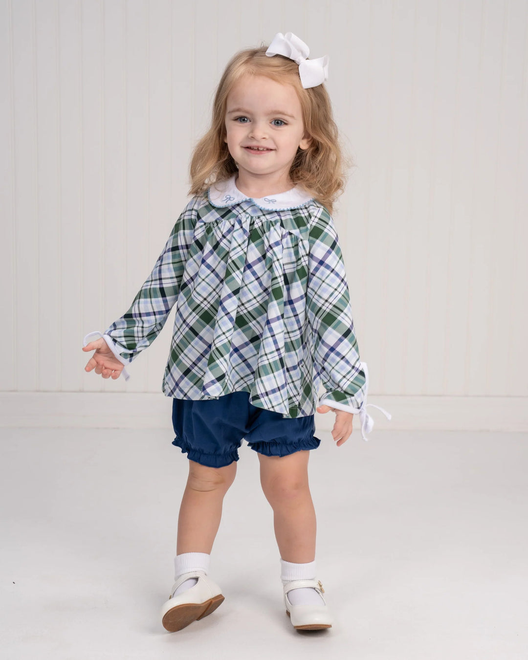 Young girl wearing a green plaid dress with a white collar and blue shorts on a light gray background