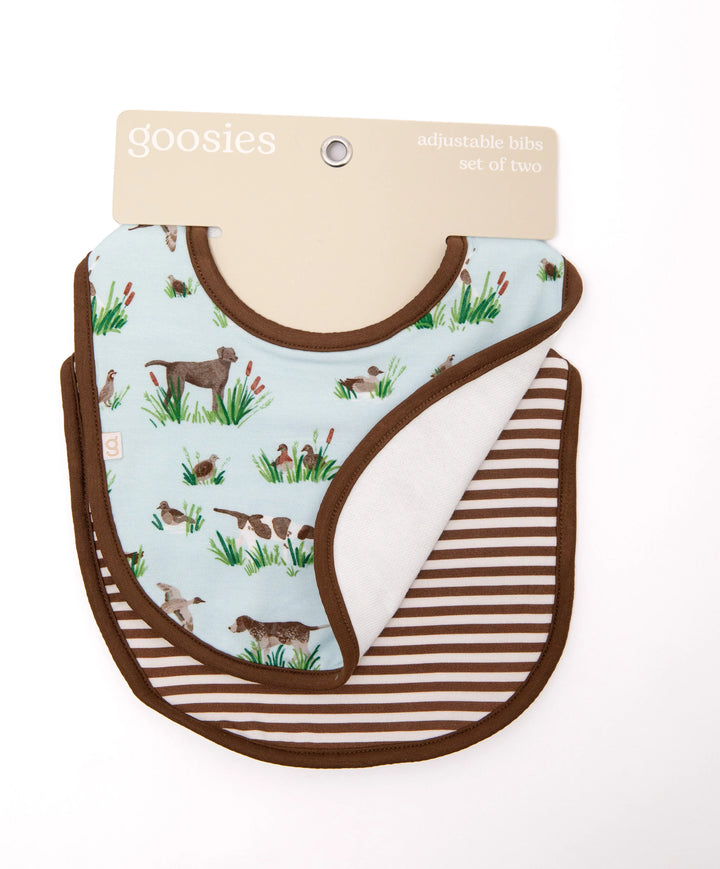 Two baby bibs from the brand Goosies, displayed on their original cardboard hanger against a white background. The front bib features a light blue background with a pattern of hunting dogs and ducks in brown and green, bordered by brown trim.