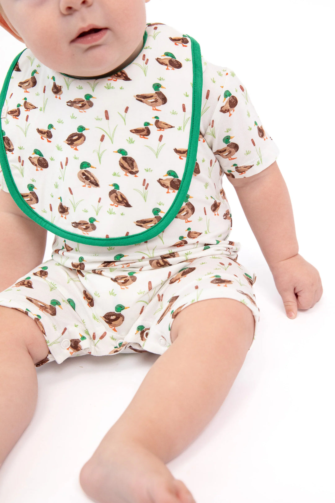 A baby wearing a short-sleeve romper and matching bib set with a brown duck print on a white background and green trim, photographed on a white studio background.