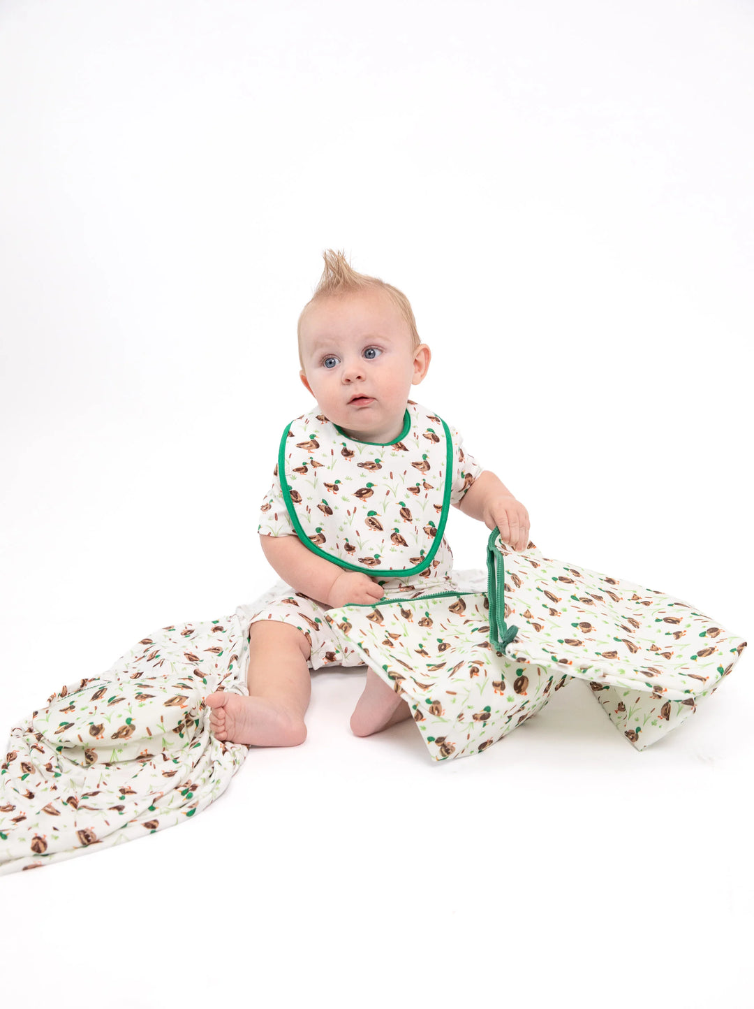 A baby sitting on a white studio background wearing a matching baby sleep sack and bib set with a brown woodland tree print and green trim.