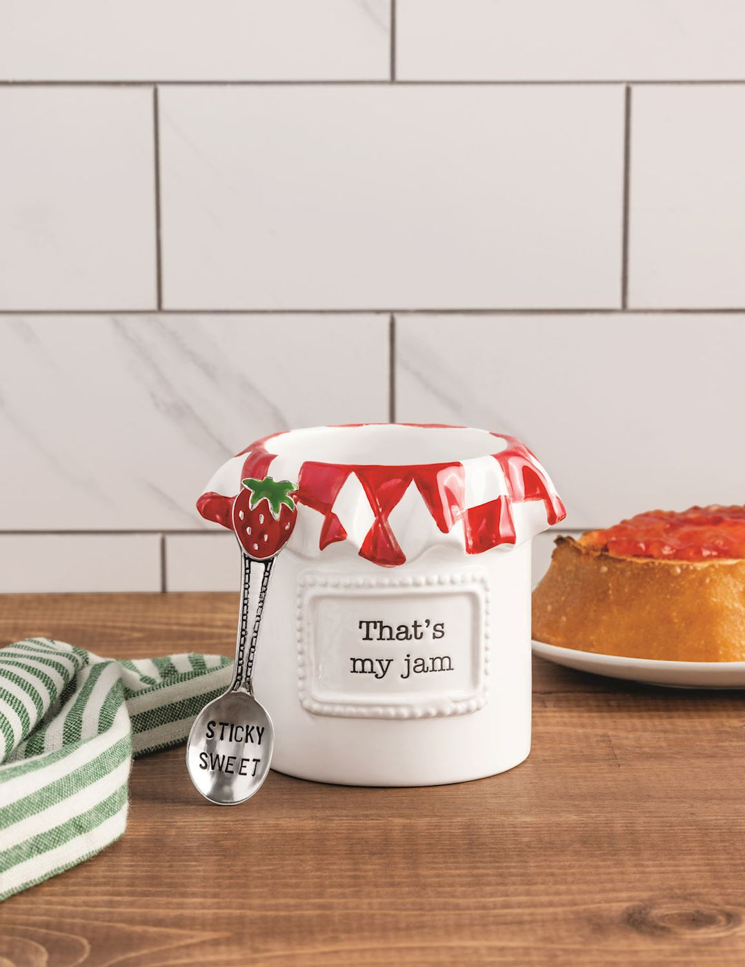 Ceramic jam jar features debossed sentiment with silver spoon on wooden table with white marble tile backspalsh