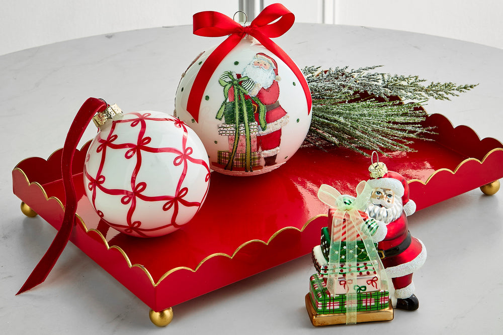 A festive holiday tabletop arrangement featuring two white Christmas ornaments with red patterns and a Santa Claus illustration, tied with red ribbons, a red scalloped tray with gold trim, green pine branches, and a small Santa gift box figurine on a light gray surface.