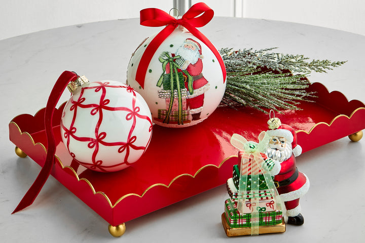 A festive holiday tabletop arrangement featuring two white Christmas ornaments with red patterns and a Santa Claus illustration, tied with red ribbons, a red scalloped tray with gold trim, green pine branches, and a small Santa gift box figurine on a light gray surface.