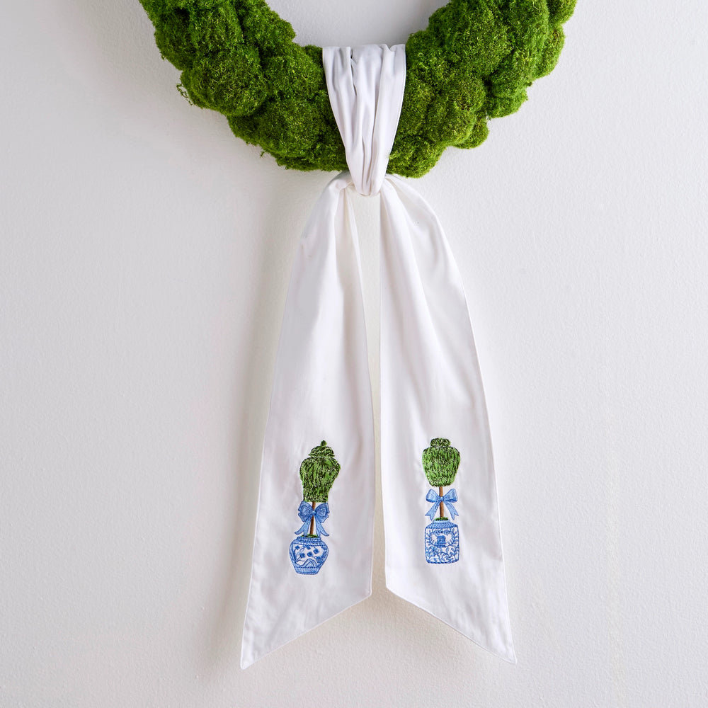 White towel with green and blue designs hanging from a green wreath on a white wall.