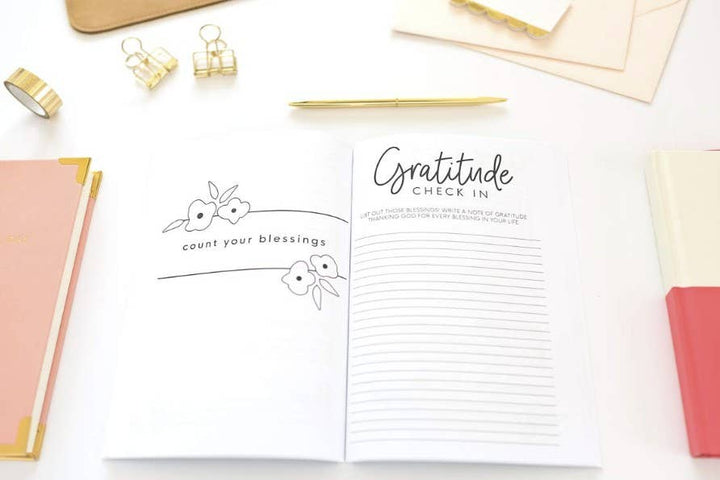 An open white gratitude journal spread on a light surface, with one page showing a bunny illustration and the phrase 'count your blessings' and the other page titled 'Gratitude CHECK IN' with lined writing space. Gold and blush stationery items are arranged around it.