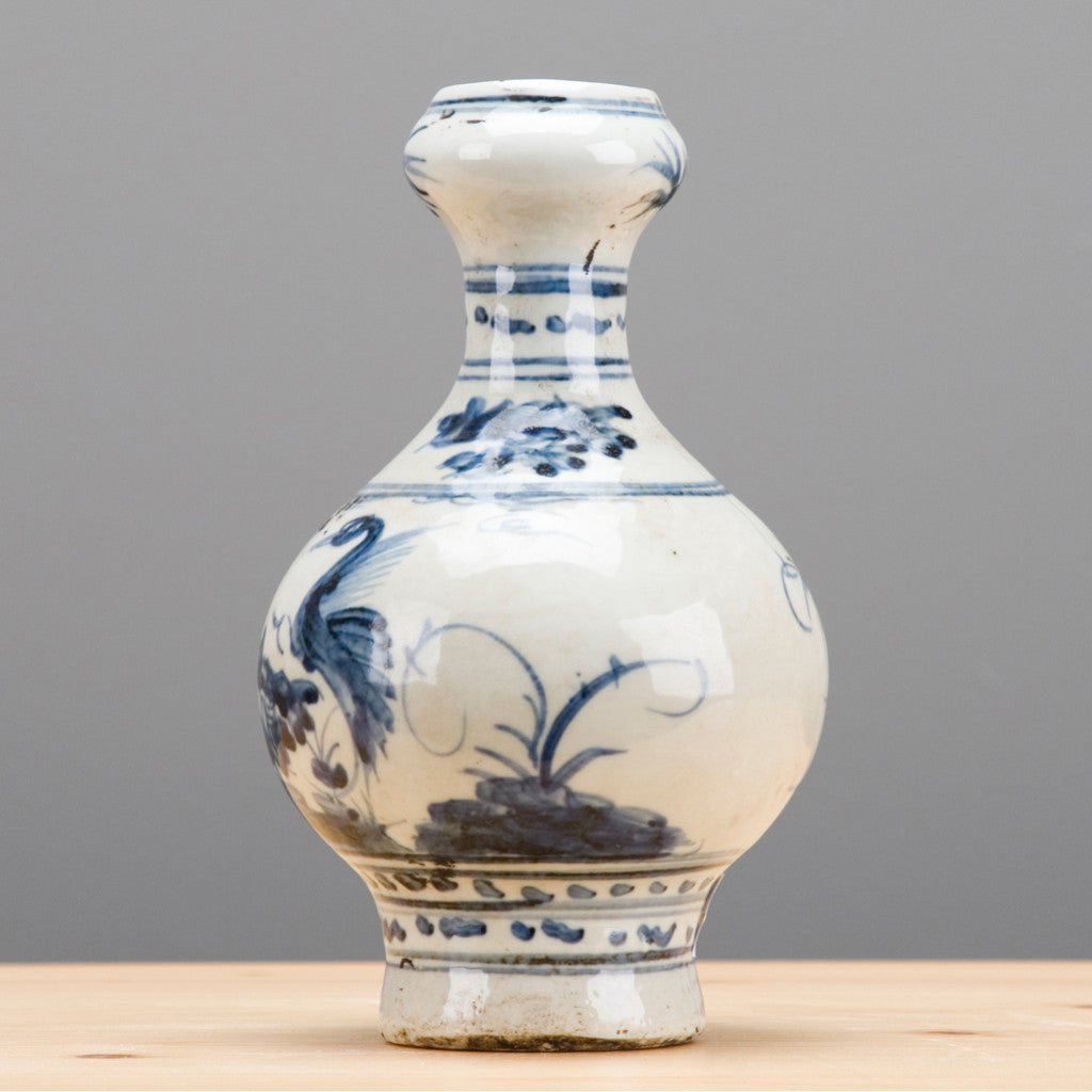 An antique-style ceramic vase with a bulbous body and narrow neck, decorated in blue and white with hand-painted floral and bird motifs, standing on a light wooden surface against a neutral gray background.