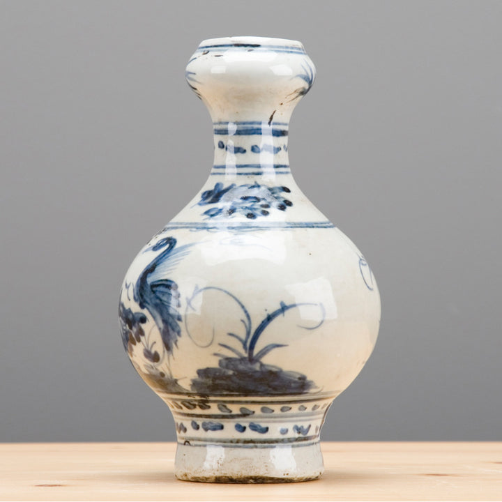 An antique-style ceramic vase with a bulbous body and narrow neck, decorated in blue and white with hand-painted floral and bird motifs, standing on a light wooden surface against a neutral gray background.
