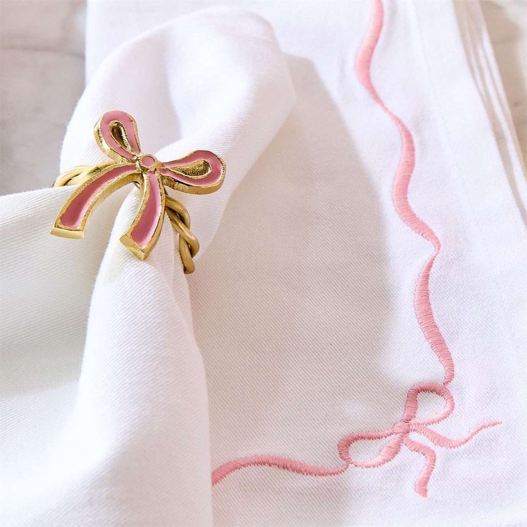 Pink and gold bow-shaped napkin ring on a white napkin with pink embroidery.