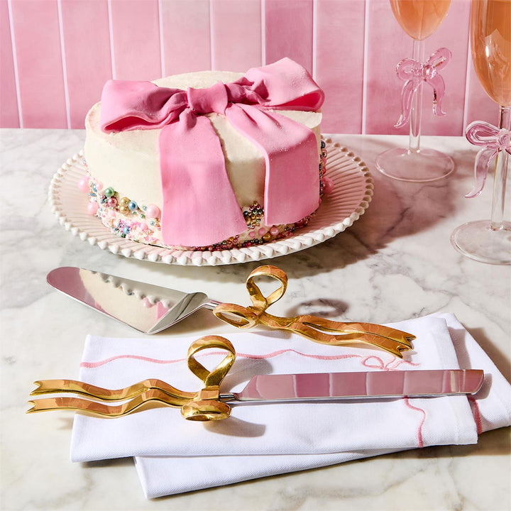 Decorative cake with pink bow, gold cake server, and pink glasses on marble surface