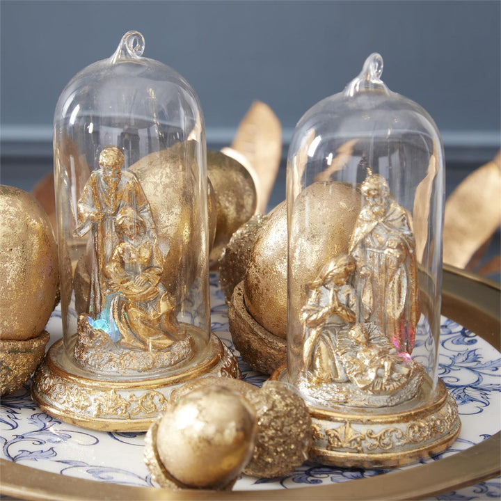 Two clear glass cloche dome ornaments, each housing a small gold-toned nativity figurine, displayed on a decorative tray with gold eggs against a dark blue-gray background.