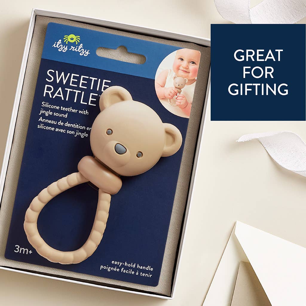 A boxed silicone baby teether rattle shown in its retail packaging on a light cream background. The product is a beige bear-shaped teether with a matching loop handle, attached to a dark blue backing card.