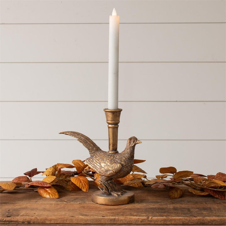 Decorative candle holder with a pheasant design on a wooden surface with leaves and a white candle.