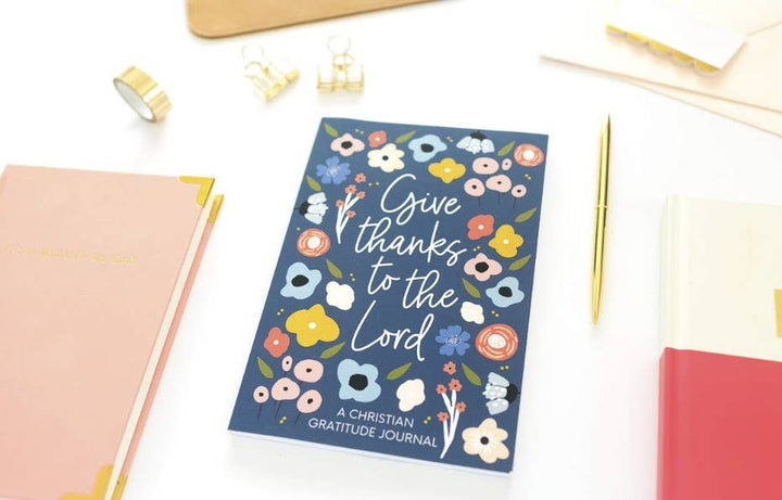 A Christian Gratitude Journal with a navy blue cover decorated with colorful flowers and the text 'Give thanks to the Lord', styled in a flat-lay on a white desk with stationery.