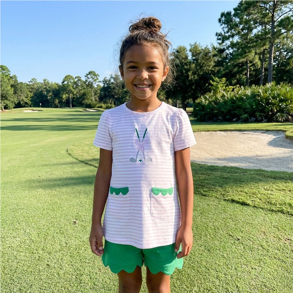 Young girl standing on a golf course wearing a pink striped t-shirt with green accents and green shorts.