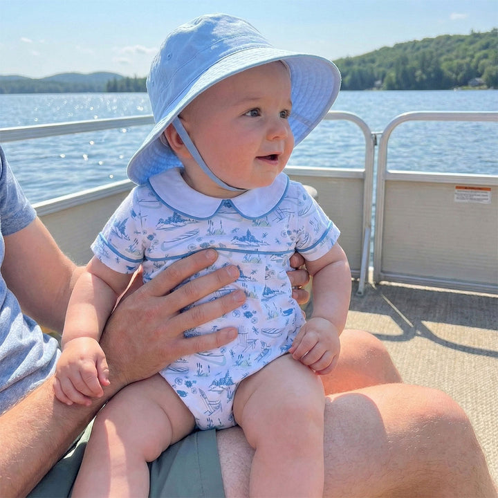 Baby in a blue hat and outfit sitting on a person's lap on a boat with water and sky in the background