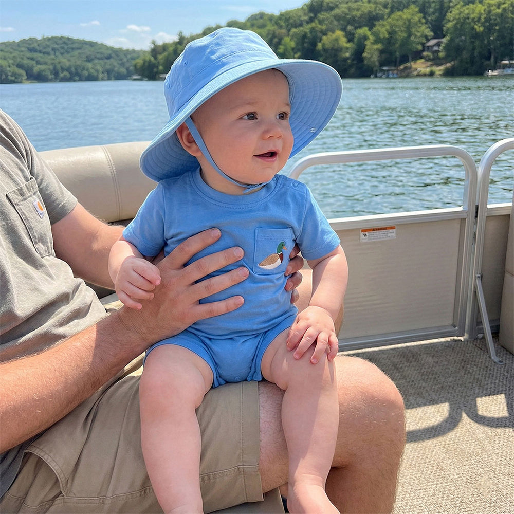 Baby in a blue outfit and hat sitting on a person's lap on a boat with a lake and trees in the background.