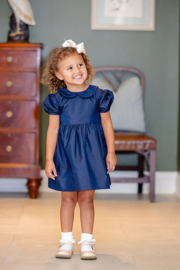 Young girl in a blue dress standing in a room with wooden furniture and a gray wall.