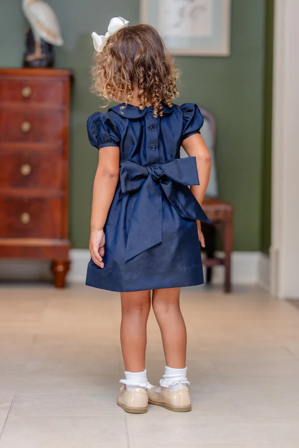 Child wearing a navy blue dress with a large bow in a room with wooden furniture and a green wall.