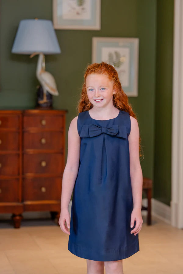 Young girl in a navy dress standing in a room with wooden furniture and framed pictures on the wall.