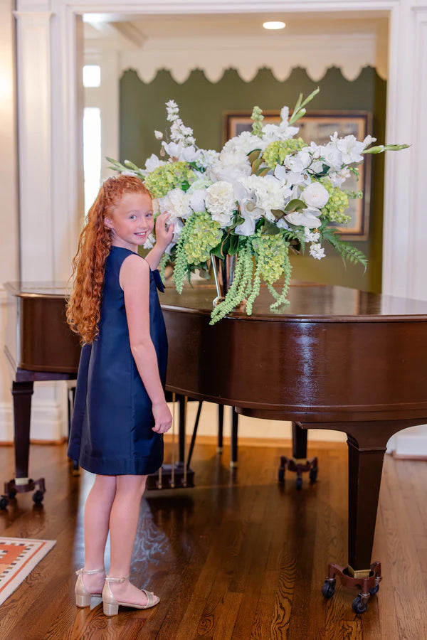 Young girl in a blue dress standing next to a grand piano with a floral arrangement.