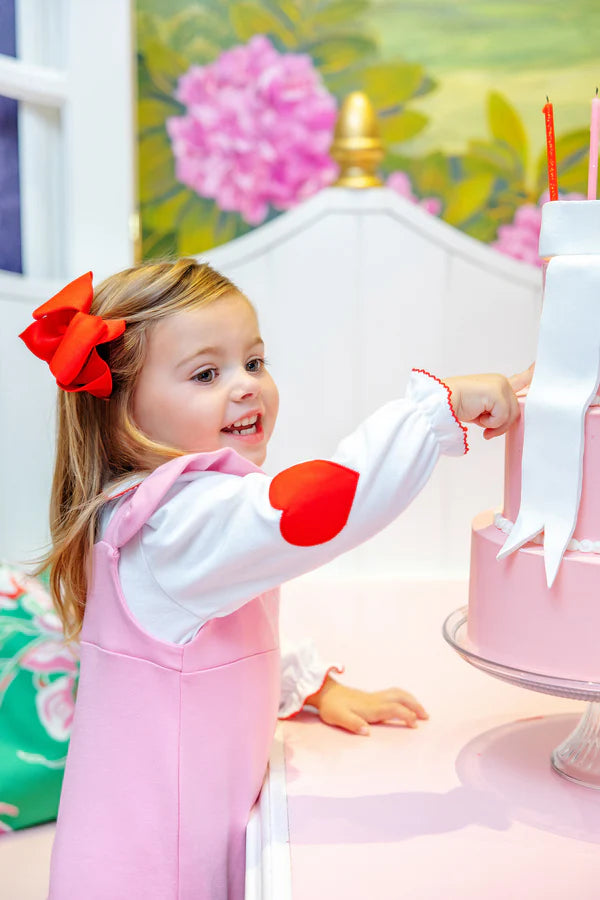 Young girl in a pink dress with a red bow, reaching towards a pink cake with white frosting.