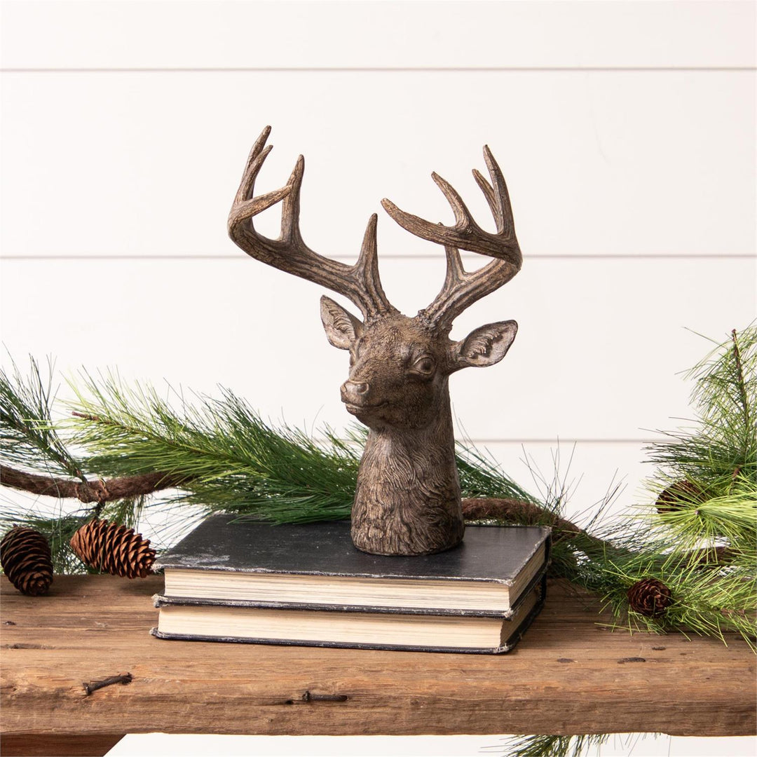Decorative deer head sculpture on books with greenery and pinecones on a wooden surface.
