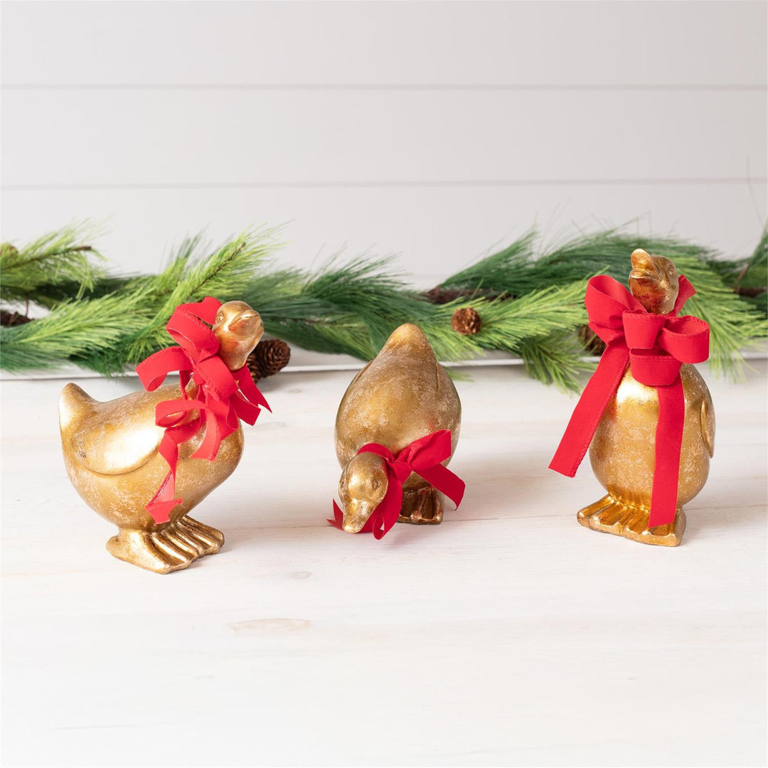 Three gold bird figurines with red bows on a white surface with greenery in the background