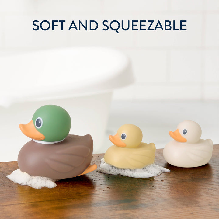 Three duck-shaped bath toys in green, yellow, and cream are lined up on a wooden surface with soap foam, with the text 'SOFT AND SQUEEZABLE' above them. A blurred white tub is in the background.