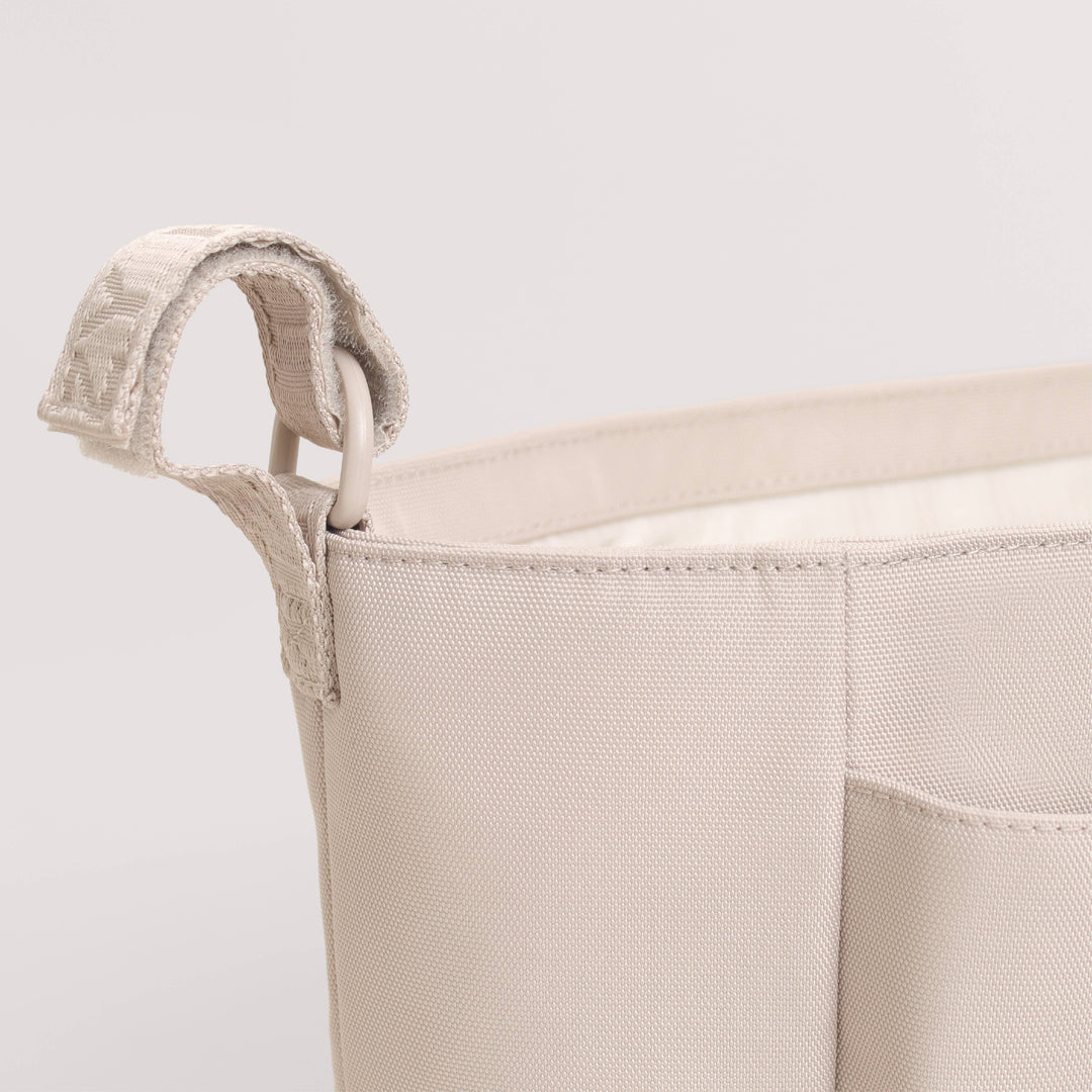 Close-up of a light beige canvas storage basket with a woven fabric handle and a front pocket, shown against a plain light gray background.