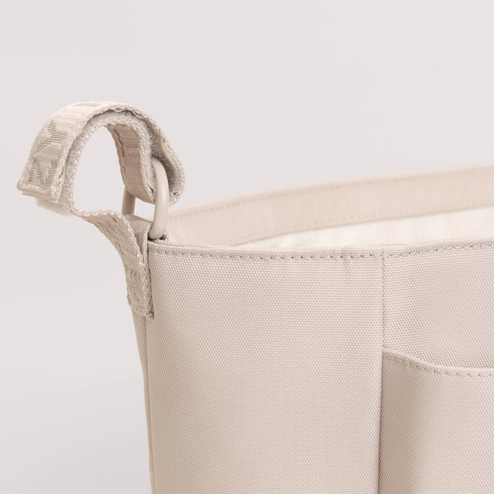 Close-up of a light beige canvas storage basket with a woven fabric handle and a front pocket, shown against a plain light gray background.