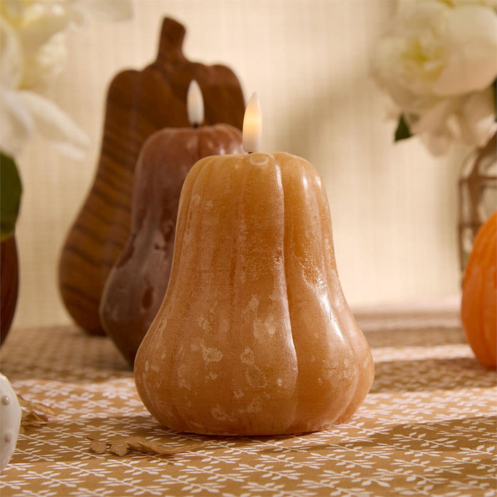 Gourd-shaped candle with a lit wick on a decorative surface with pumpkins and flowers.