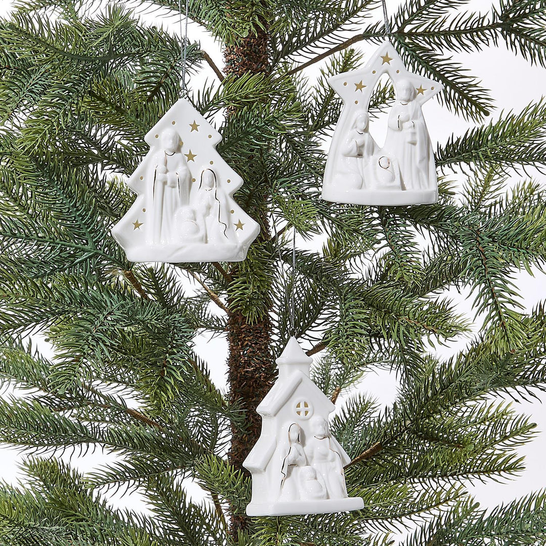 White nativity scene ornaments hanging on a Christmas tree.