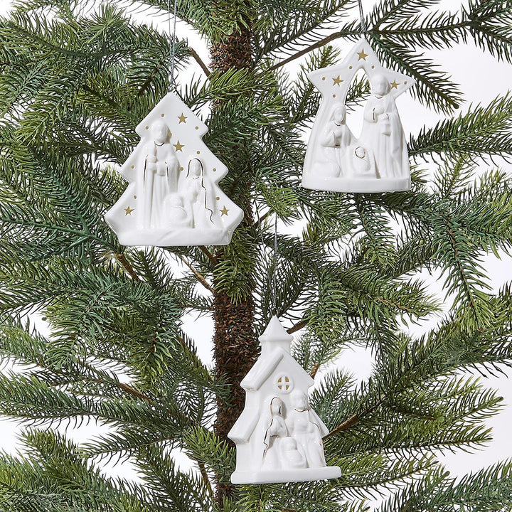 White nativity scene ornaments hanging on a Christmas tree.