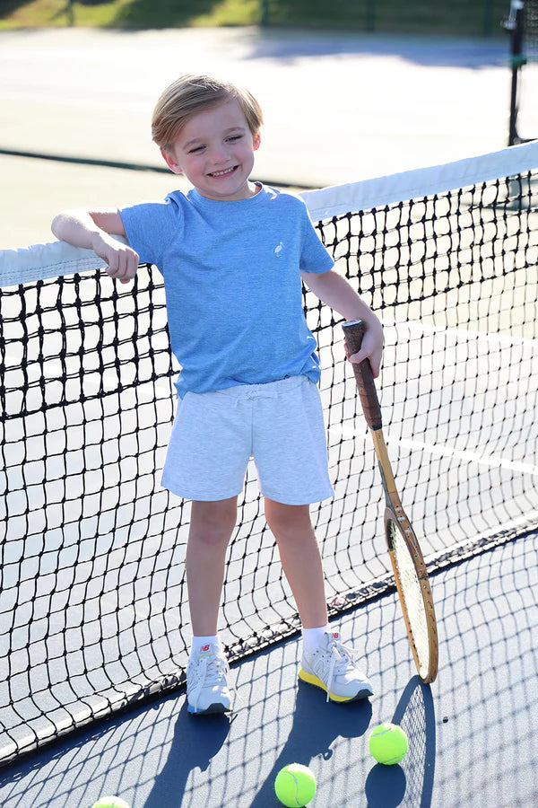 Child in blue shirt and white shorts standing on a tennis court with a racket and ball.