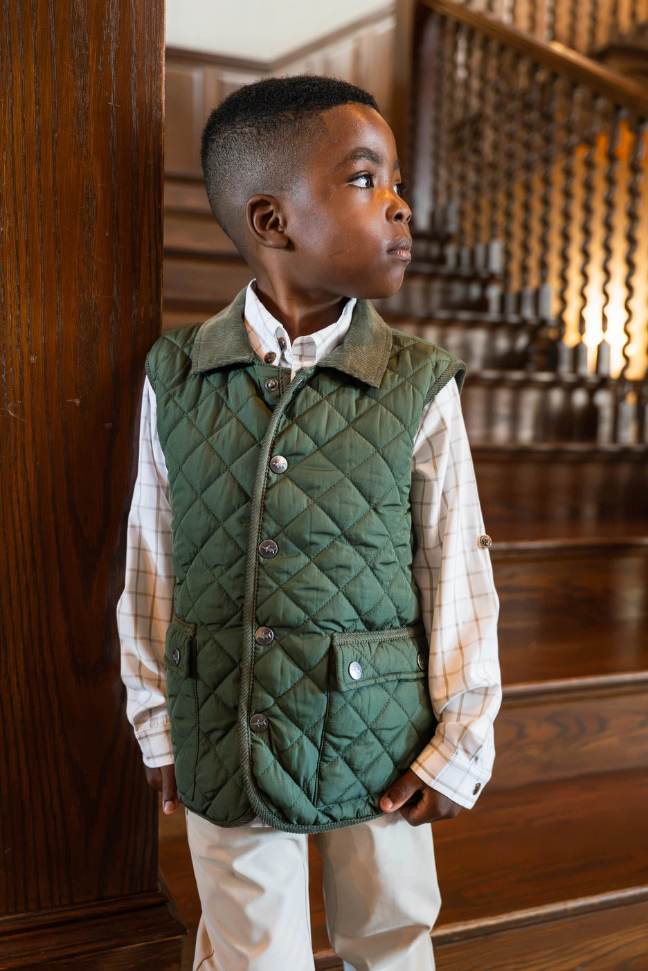close up of little boy wearing green quilted vest in front of wooden staircase