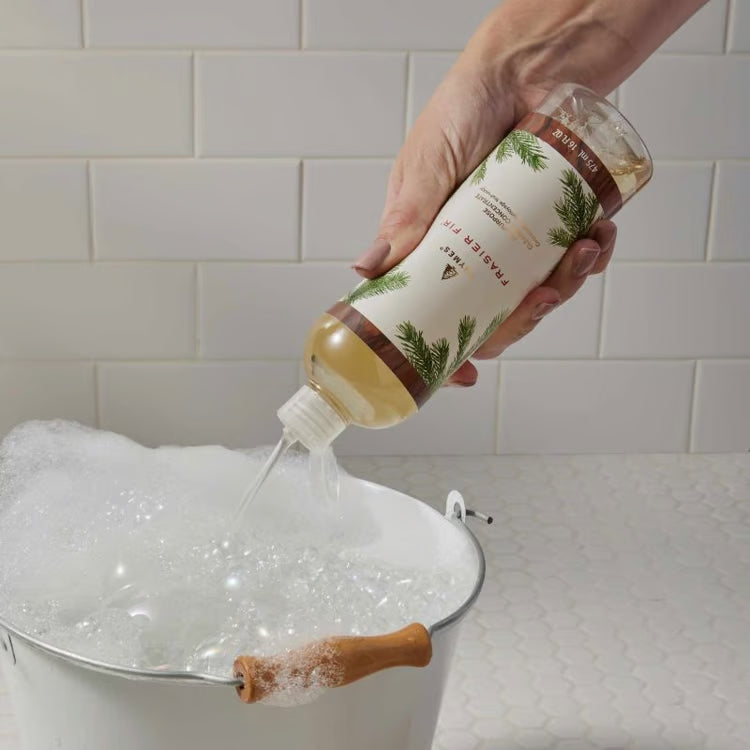 A hand holds a bottle of bath salts and pours the clear liquid into a white tub filled with foam, set against a white subway tile wall.